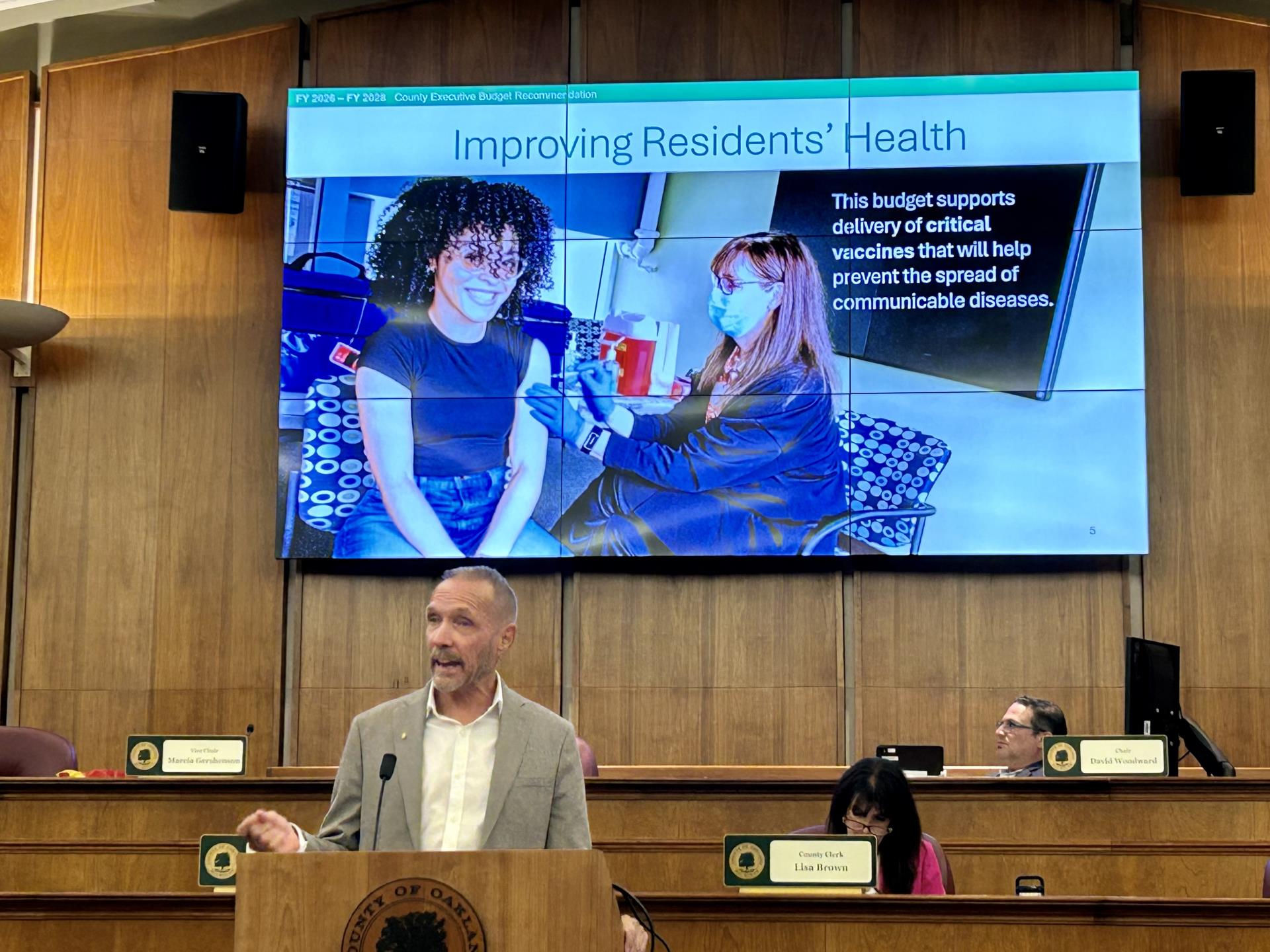 Man stands behind a podium in front of a screen that depicts vaccination